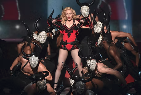 Madonna performing on stage in a red and black horned costume, surrounded by dancers wearing horned masks